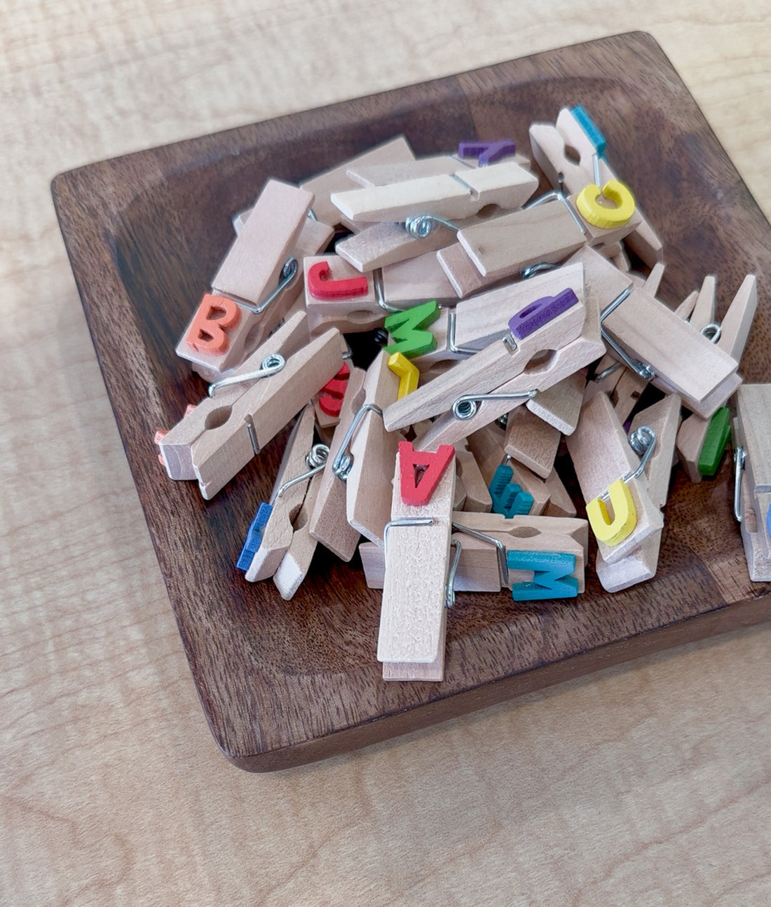 A pile of wooden clothespins with colorful alphabet letters attached, sitting in a square wooden tray for letter recognition activities.