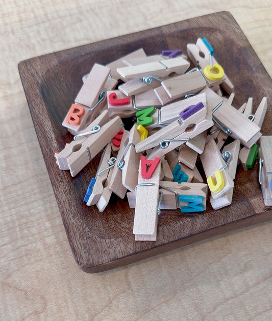 A pile of wooden clothespins with colorful alphabet letters attached, sitting in a square wooden tray for letter recognition activities.